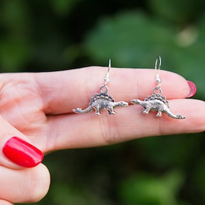 Op de afbeelding: Een paar zilveren dinosaurus oorbellen. De oorbellen zijn gevormd als een stegosaurus en hebben een hangend ontwerp.