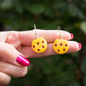 May include: A pair of dangle earrings featuring a yellow cookie with brown chocolate chips. The earrings are made of polymer clay and have silver hooks.