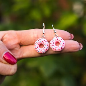 Könnte beinhalten: Ein Paar rosafarbene Donut-Ohrringe mit bunten Streuseln. Die Ohrringe sind aus Polymer Clay gefertigt und haben silberne Haken.