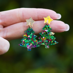 以下が含まれることがあります： クリスマスツリーのイヤリングのペア。各イヤリングは、カラフルなグリッターアクセントが付いた緑の木の形をしています。各木の上には金の星があります。イヤリングは、ぼやけた緑の背景に対して表示されています。