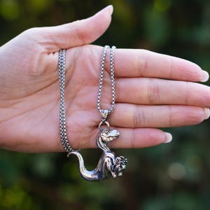 Puede incluir: Un collar con colgante de dinosaurio plateado. El colgante es una representación detallada en tres dimensiones de un dinosaurio. La cadena del collar es de color plateado a juego. El fondo es verde borroso.