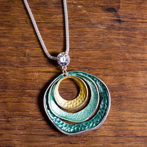 Puede incluir: Collar plateado con un colgante de tres círculos texturizados superpuestos en verde y amarillo. El collar se exhibe sobre una superficie de madera, mostrando el diseño y el contraste de color de la joya.