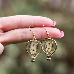 Può includere: Orecchini color oro a forma di mongolfiera. Ogni orecchino presenta un involucro in resina trasparente con fiori bianchi e un gancio color oro. Gli orecchini sono tenuti da una mano con smalto rosso.