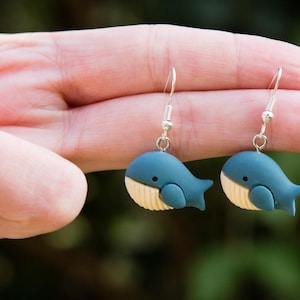 Può includere: Un paio di orecchini a forma di balena blu con pancia color crema e piccoli occhi neri, appesi a ganci color argento. Gli orecchini sono tenuti da una mano con lo smalto rosso. Un accessorio originale.