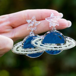 Op de afbeelding: Een paar oorbellen met ruimtethema. Elke oorbel heeft een blauw planeetontwerp met een witte ring en een heldere stervormige top. De oorbellen worden tegen een vage groene achtergrond gehouden.