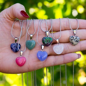 Könnte beinhalten: Eine Sammlung herzförmiger Edelsteinanhänger an Silberketten. Die Anhänger sind in verschiedenen Farben erhältlich, darunter Rosa, Blau, Grün und Weiß. Die Halsketten werden in einer Hand vor einem verschwommenen grünen Hintergrund gehalten.
