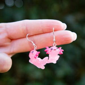 Può includere: Un paio di orecchini a forma di axolotl rosa con ganci argentati. Gli axolotl hanno facce sorridenti e branchie rosa. Un accessorio carino e stravagante.