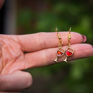 Op de afbeelding: Goudkleurige oorhangers met een wijnglasontwerp. Elke oorbel heeft een gouden frame met een rode emaille vulling, die lijkt op een glas rode wijn. De oorbellen zijn bevestigd aan goudkleurige oorhaken.
