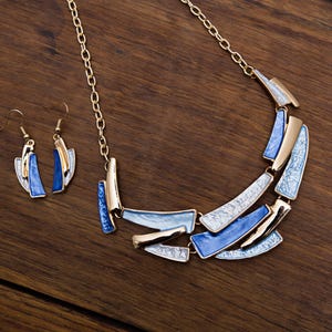 Puede incluir: Un conjunto de collar y pendientes a juego en tono dorado. El collar presenta una serie de formas geométricas azules, blancas y doradas. Los pendientes reflejan el diseño. Las joyas se exhiben sobre una superficie de madera.