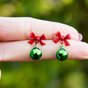 Op de afbeelding: Een paar kerstoorbellen. Elke oorbel heeft een rode strik en een groene belvormige bedel. De oorbellen zijn klein en delicaat, met een glanzende afwerking. De achtergrond is een vage groene kleur.