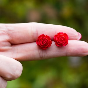 Puede incluir: Un par de pendientes de botón con forma de rosa roja brillante. Los pendientes son pequeños, con pétalos detallados, y se muestran en la mano de una persona. El fondo está borroso, con tonos verdes y marrones.