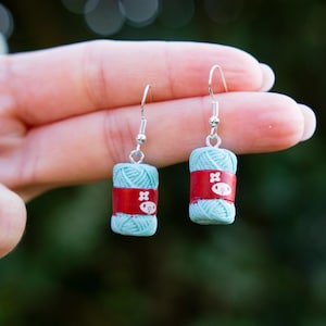 May include: A pair of handmade earrings featuring miniature yarn balls. Each earring has a light blue yarn ball wrapped with a red band. The red band has a white graphic. The earrings hang from silver-colored fish hook ear wires.
