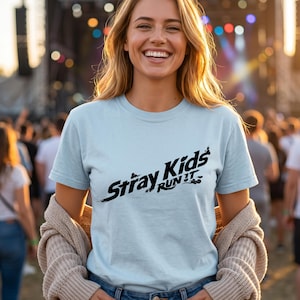 May include: A light blue short-sleeved t-shirt featuring the black text Stray Kids RUN IT printed across the chest. The shirt has a crew neckline and a standard fit, showcasing a graphic design with a distressed, brush-stroke style font.