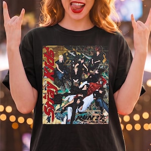 May include: A black short-sleeved t-shirt featuring a graphic print of the music group Stray Kids. The design includes the red text Stray Kids vertically on the left and the text MANIAC WORLD TOUR at the bottom, over a collage of band members.