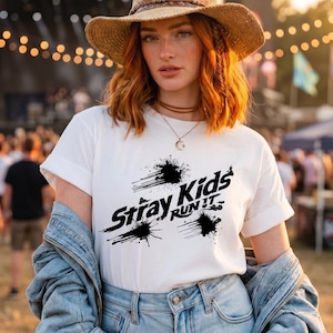 May include: A white short-sleeved t-shirt featuring a black graphic print that reads Stray Kids RUN IT, accented with abstract ink splatter designs. The shirt is worn with a light blue denim jacket, a straw hat, and a delicate gold crescent moon necklace.