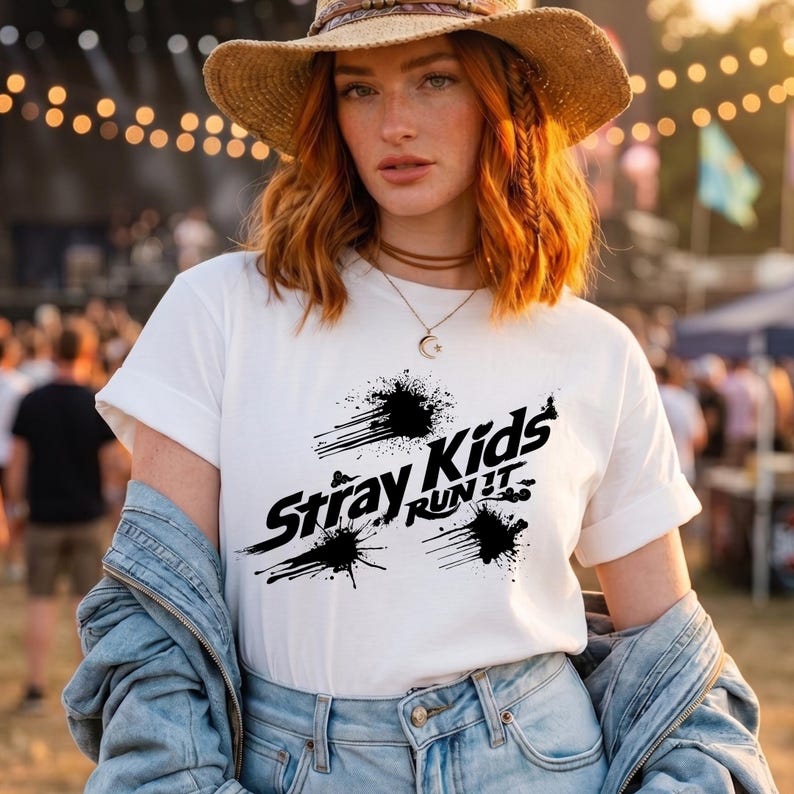 May include: A white short-sleeved t-shirt featuring a black graphic print that reads Stray Kids RUN IT, accented with abstract ink splatter designs. The shirt is worn with a light blue denim jacket, a straw hat, and a delicate gold crescent moon necklace.