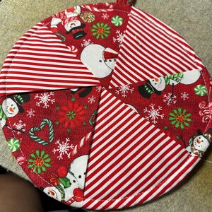 Puede incluir: Un salvamanteles navideño redondo y acolchado con un lazo para colgar. El salvamanteles presenta un diseño de patchwork con tela a rayas rojas y blancas y tela roja con muñecos de nieve, copos de nieve y patrones de bastones de caramelo. El salvamanteles mide aproximadamente 20 cm de diámetro.