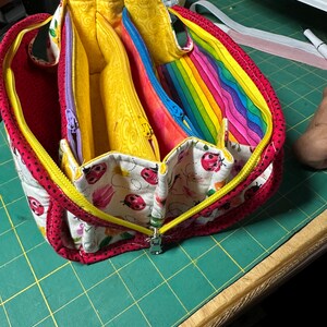May include: A red, zippered organiser with multiple compartments. The interior features yellow, pink, and blue zippered sections. The exterior has a ladybug and floral pattern on a white background, with a yellow zip and red trim. A rainbow fabric is visible inside.
