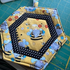 May include: A yellow hexagon-shaped pot holder with a black and white polka dot border. The pot holder features a bee and flower design with a blue and gray bee character. The pot holder has a loop handle.