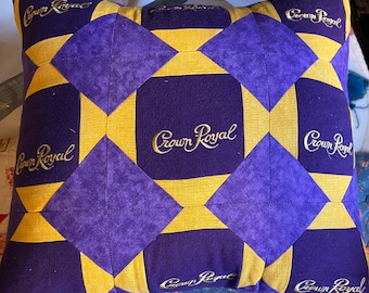 Op de afbeelding: Een paars en goud gestikt kussen met de woorden "Crown Royal" geborduurd op elk paneel.