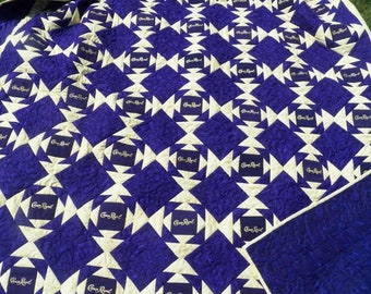 Op de afbeelding: Een paarse en witte quilt met een herhalend patroon van Crown Royal-logo's. De quilt is gemaakt van vierkante stoffen met een witte achtergrond en een paars vierkant in het midden met het Crown Royal-logo.