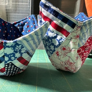 Puede incluir: Dos salvabols de cuenco acolchados con patrones patrióticos. Uno presenta un fondo azul con lunares blancos, mientras que el otro muestra un diseño de patchwork con telas rojas, blancas y azules, incluyendo estrellas y rayas.