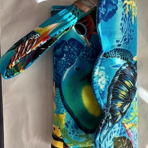 Könnte beinhalten: Eine blaue Stoffgeldbörse mit Handschlaufe, die ein lebendiges Wassermotiv aufweist. Die Geldbörse zeigt ein farbenfrohes Muster aus Fischen, Korallen und anderen Meereslebewesen. Ein silberfarbener Schlüsselring ist an der Geldbörse befestigt.