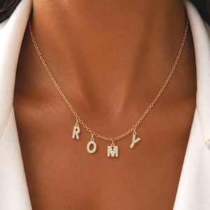 Peut inclure: Un collier à chaîne dorée orné de quatre breloques en forme de lettres pendantes formant le nom ROMY. Chaque breloque est incrustée de petites pierres claires et scintillantes qui captent la lumière, suspendues à une fine chaîne à maillons.