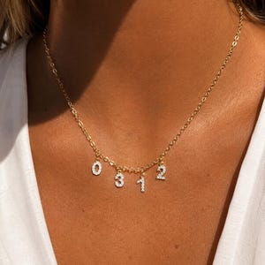 Op de afbeelding: Een goudkleurige ketting met vier bungelende bedels in de vorm van de cijfers 0, 3, 1 en 2. Elke cijferbedel is ingelegd met kleine, heldere, fonkelende steentjes die het licht vangen, hangend aan een fijne schakelketting.