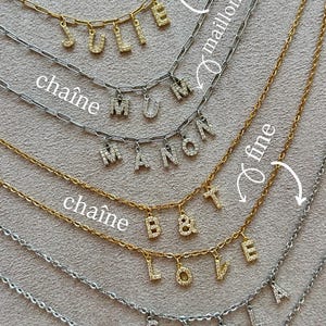 May include: A collection of gold and silver chain necklaces with pendant letters spelling out names. The letters are encrusted with small, sparkling stones. The necklaces are displayed on a neutral-toned surface.
