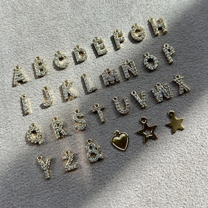 May include: A collection of gold-colored alphabet charms, each encrusted with small, clear crystals. The letters A through Z are displayed, along with a heart and star charm. The charms are arranged on a light gray surface.
