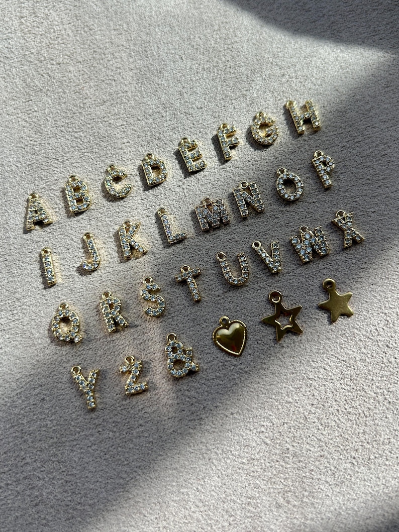 May include: A collection of gold-colored alphabet charms, each encrusted with small, clear crystals. The letters A through Z are displayed, along with a heart and star charm. The charms are arranged on a light gray surface.