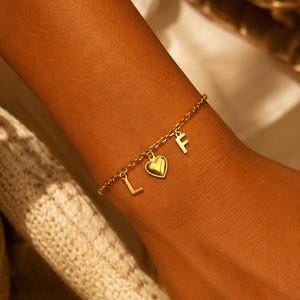 Puede incluir: Una pulsera de cadena dorada con tres dijes colgantes: una letra mayúscula L, un corazón pequeño con borde de cuentas y una letra mayúscula F. Los delicados eslabones de la cadena de metal conectan cada dije en un accesorio sencillo.