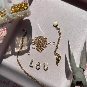 May include: A jewelry-making scene with gold-colored chain, jump rings, and the letters "LOU" spelled out in small, gem-studded letters. Pliers are visible, along with small plastic containers of gold jump rings. The items are arranged on a pink surface.