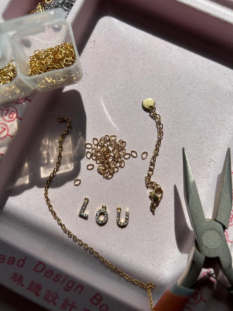 May include: A jewelry-making scene with gold-colored chain, jump rings, and the letters "LOU" spelled out in small, gem-studded letters. Pliers are visible, along with small plastic containers of gold jump rings. The items are arranged on a pink surface.