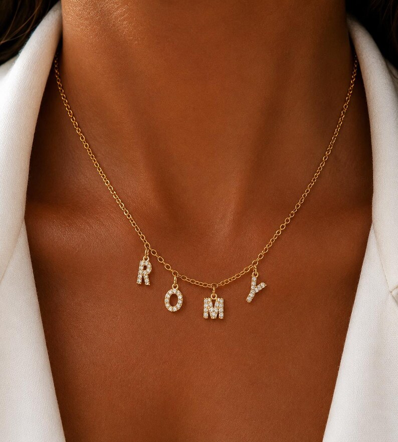 May include: A gold-colored necklace with the letters R, O, M, and Y spelled out in individual, sparkling, diamond-like letters. The necklace is worn around the neck and hangs against a neutral background.