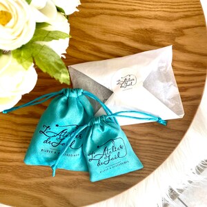 May include: Two turquoise drawstring pouches and a white envelope with a sticker, all on a wooden tray. The pouches have the text "L'Atelier de Ja&euml;l BIJOUX & ACCESSOIRES" printed on them. White flowers are in the background.