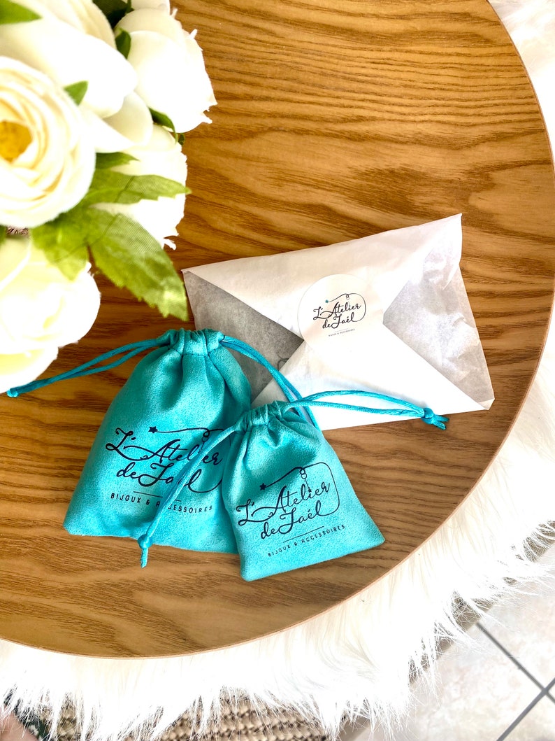 May include: Two turquoise drawstring pouches and a white envelope with a sticker, all on a wooden tray. The pouches have the text "L'Atelier de Ja&euml;l BIJOUX & ACCESSOIRES" printed on them. White flowers are in the background.