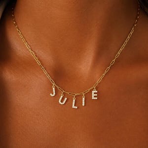 Pode incluir: Um colar de corrente dourada com cinco pingentes de letras pendentes que formam o nome JULIE. Cada letra é incrustada com pequenas pedras claras e brilhantes, e a corrente consiste em elos ovais alongados com um acabamento dourado uniforme.