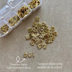 May include: Gold-colored letter charms with clear rhinestones, scattered on a beige surface. Some letters are in clear plastic containers. The word "OUI" is spelled out with the charms. The approximate letter height is 8mm.