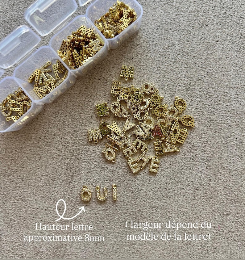 May include: Gold-colored letter charms with clear rhinestones, scattered on a beige surface. Some letters are in clear plastic containers. The word "OUI" is spelled out with the charms. The approximate letter height is 8mm.
