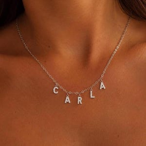 May include: Silver necklace with the name "CARLA" spelled out in individual, sparkling letter charms. The necklace is worn around the neck, with the letters hanging below the chain.
