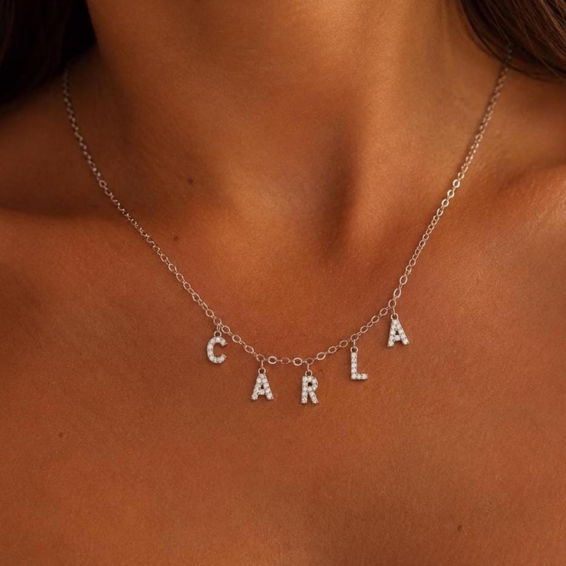 May include: Silver necklace with the name "CARLA" spelled out in individual, sparkling letter charms. The necklace is worn around the neck, with the letters hanging below the chain.