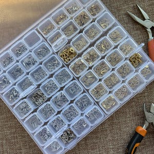May include: A clear plastic organizer filled with small, ornate metal embellishments in various shapes and finishes. The compartments hold gold and silver-toned filigree pieces. Pliers with orange and black handles are visible on the side, suggesting jewelry making.