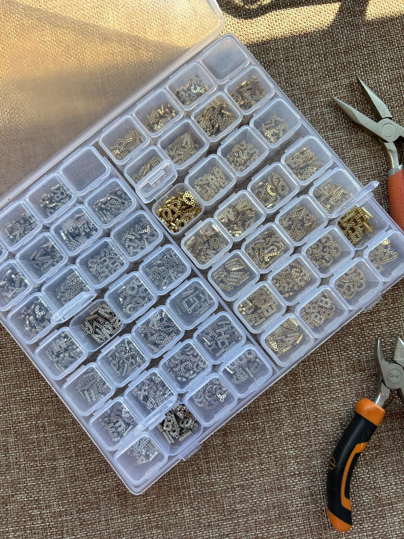 May include: A clear plastic organizer filled with small, ornate metal embellishments in various shapes and finishes. The compartments hold gold and silver-toned filigree pieces. Pliers with orange and black handles are visible on the side, suggesting jewelry making.