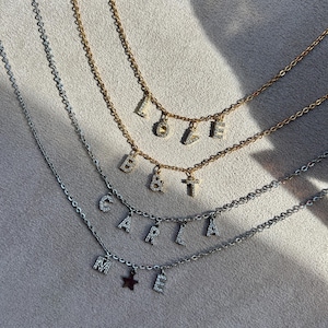 May include: Three delicate necklaces with letter pendants. One gold-toned necklace spells "LOVE", another spells "B&T", and a silver-toned necklace spells "CARLA" with a star charm and the letter "E".