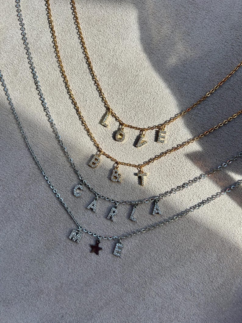 May include: Three delicate necklaces with letter pendants. One gold-toned necklace spells "LOVE", another spells "B&T", and a silver-toned necklace spells "CARLA" with a star charm and the letter "E".