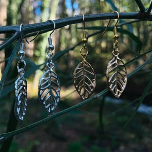 Puede incluir: Cuatro pares de pendientes con forma de hoja. Dos pares son plateados y dos dorados. Los pendientes cuelgan de una rama verde sobre un fondo borroso verde y azul.