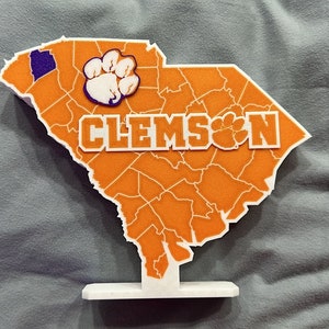 Könnte beinhalten: Orangefarbener und weißer 3D-gedruckter Kartenausschnitt von South Carolina mit dem Wort "CLEMSON" und einem Pfotenabdruck. Die Karte steht auf einem weißen Sockel.