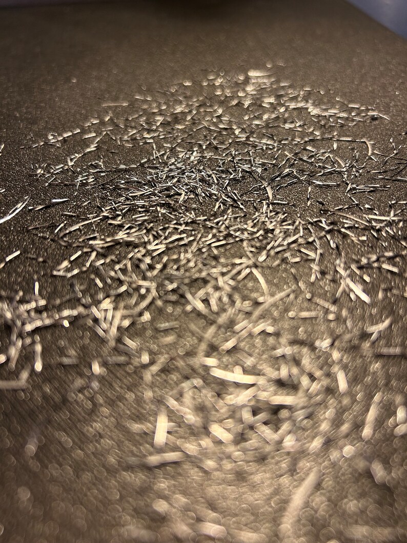 May include: A close-up image of a metallic surface covered in a large amount of silver glitter. The glitter is scattered randomly across the surface, creating a shimmering effect.